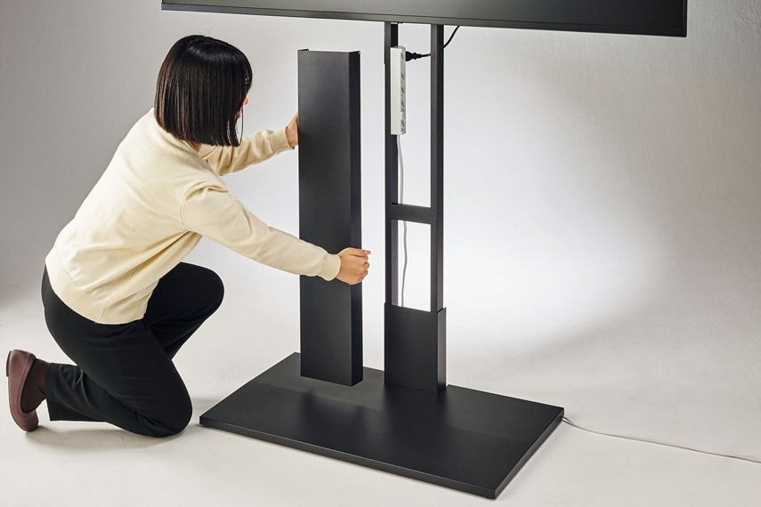 ディスプレイスタンド本体の脚部分（ディスプレイの真下）にOAタップがマグネットでくっついており、取り外し可能な前面パネルを持った人が左側にしゃがんでいる様子。