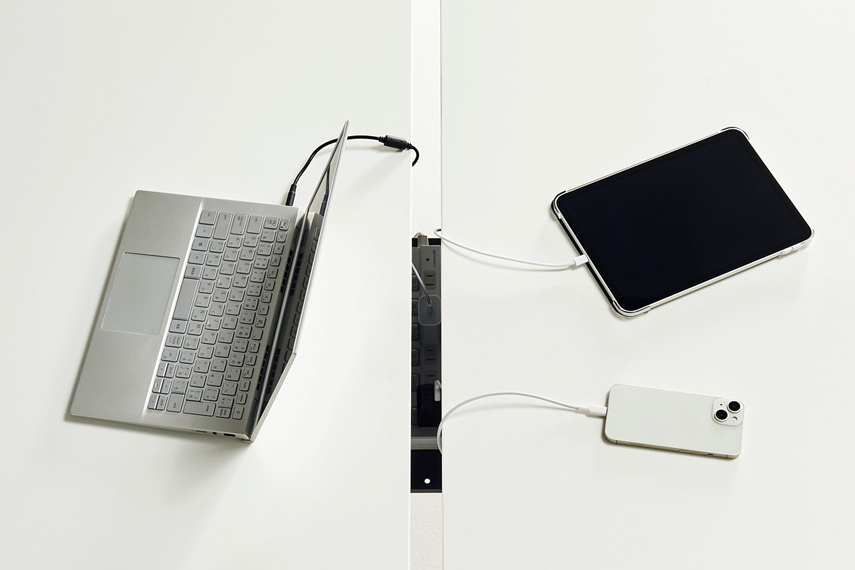 ワークテーブルの「両面タイプ」は2枚の天板で構成されており、その天板の間から配線処理ができるように設計されていることを説明する画像。ノートPCタブレット端末、スマートフォンを充電している。