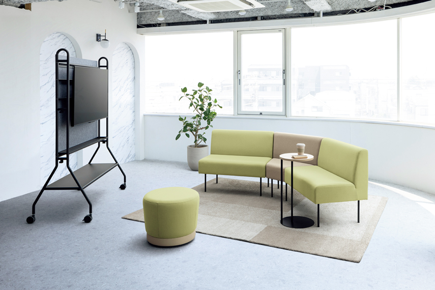 オフィスのオープン空間をイメージした場所に、ソファーが配置されている画像。2台の一般的な形状のソファーの間に「台形タイプ」のソファーを挟むことで、ゆるやかなカーブ状にレイアウトされている。ソファーの前にはディスプレイスタンドが置いてあり、ソファーの角度によるディスプレイの見やすさを表している。